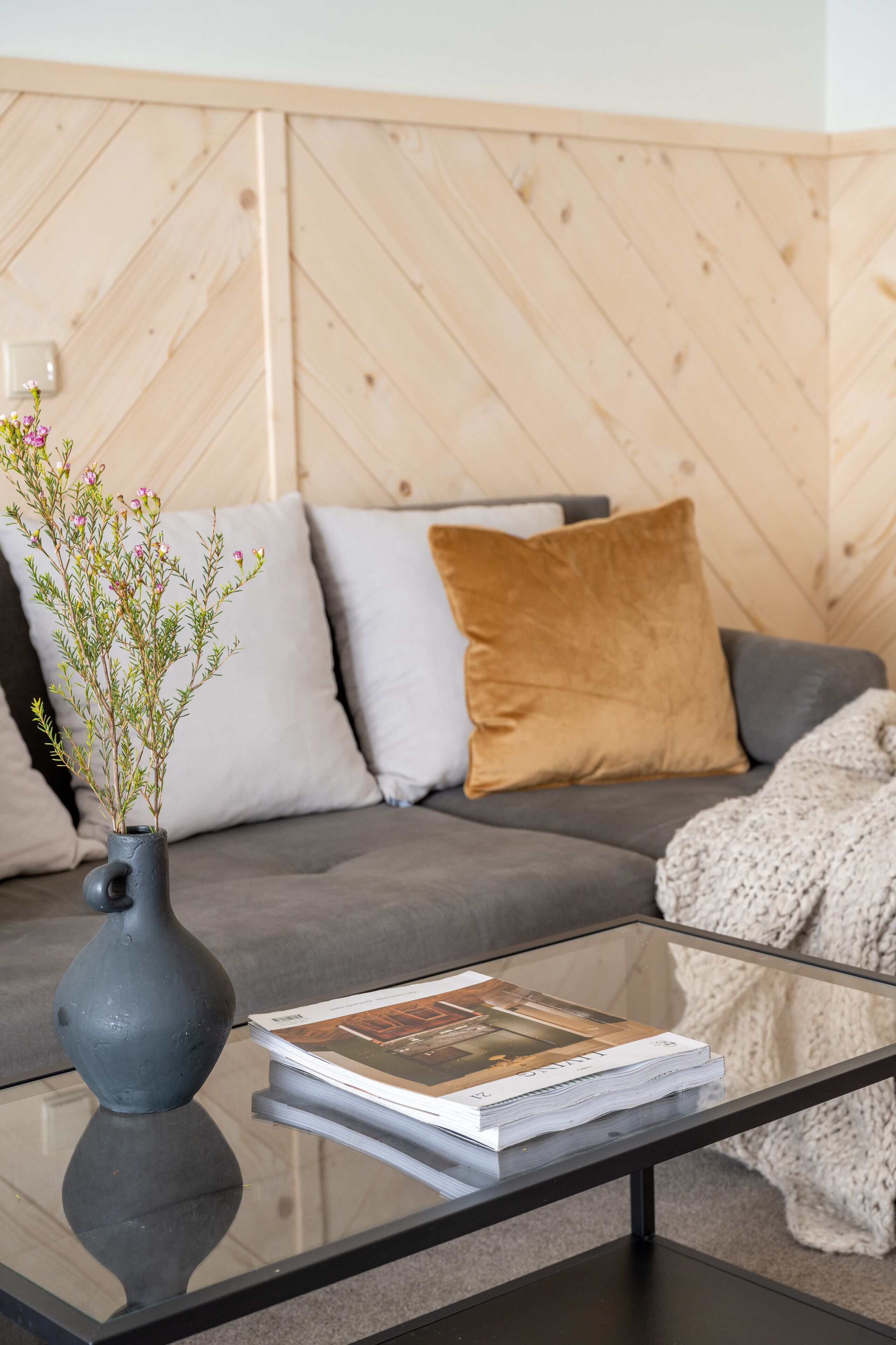 Un salon moderne avec une table basse en verre, un canapé gris et un lambris mural en bois.