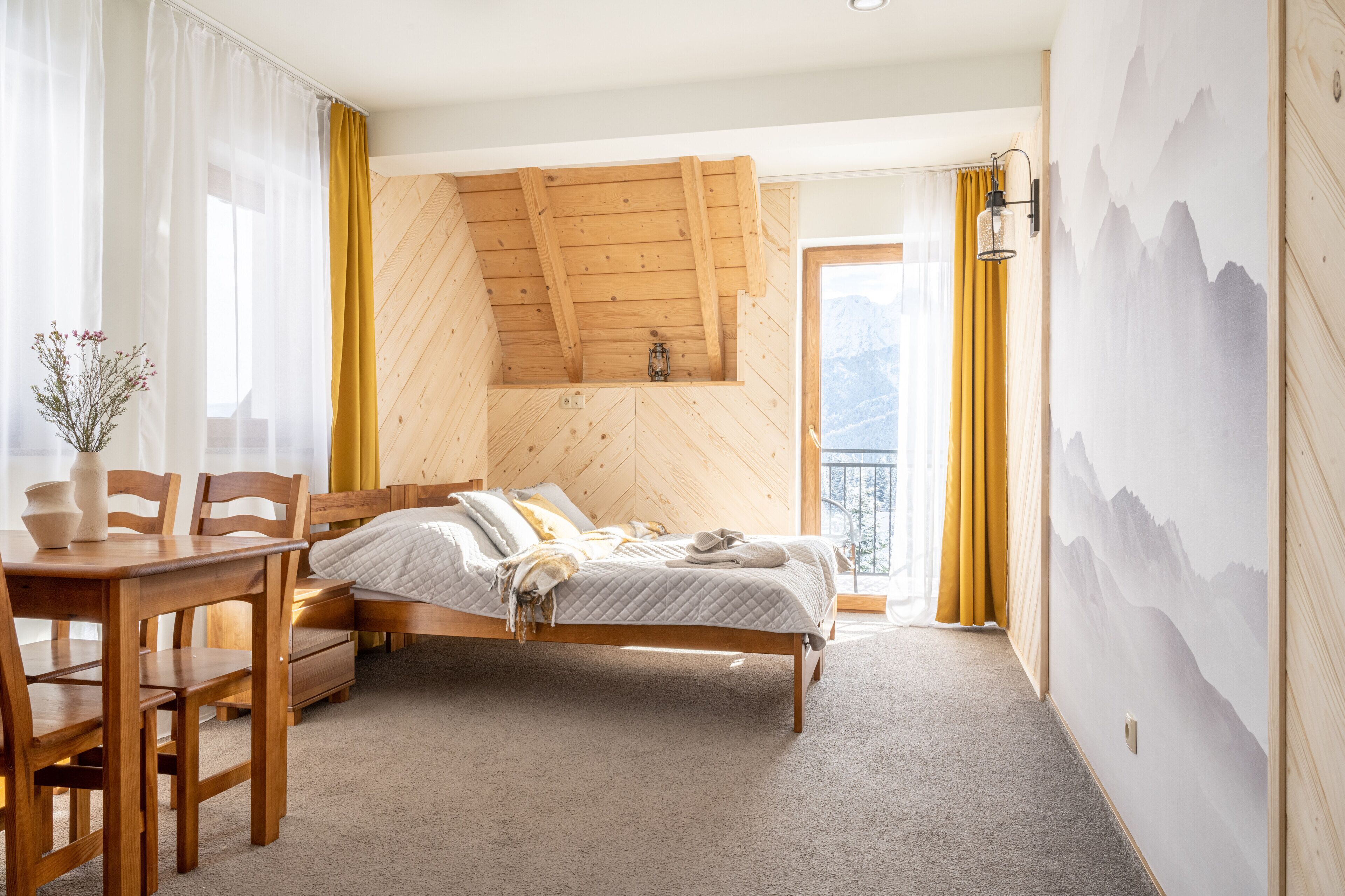 A bedroom with a wooden ceiling, a bed, a table with chairs, and a view of mountains through the window.