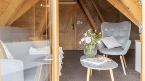 Een gezellig slaapkamer op zolder met een grijs gestoffeerde fauteuil, een klein rond tafeltje met een bloevas en een net opgemaakt bed.