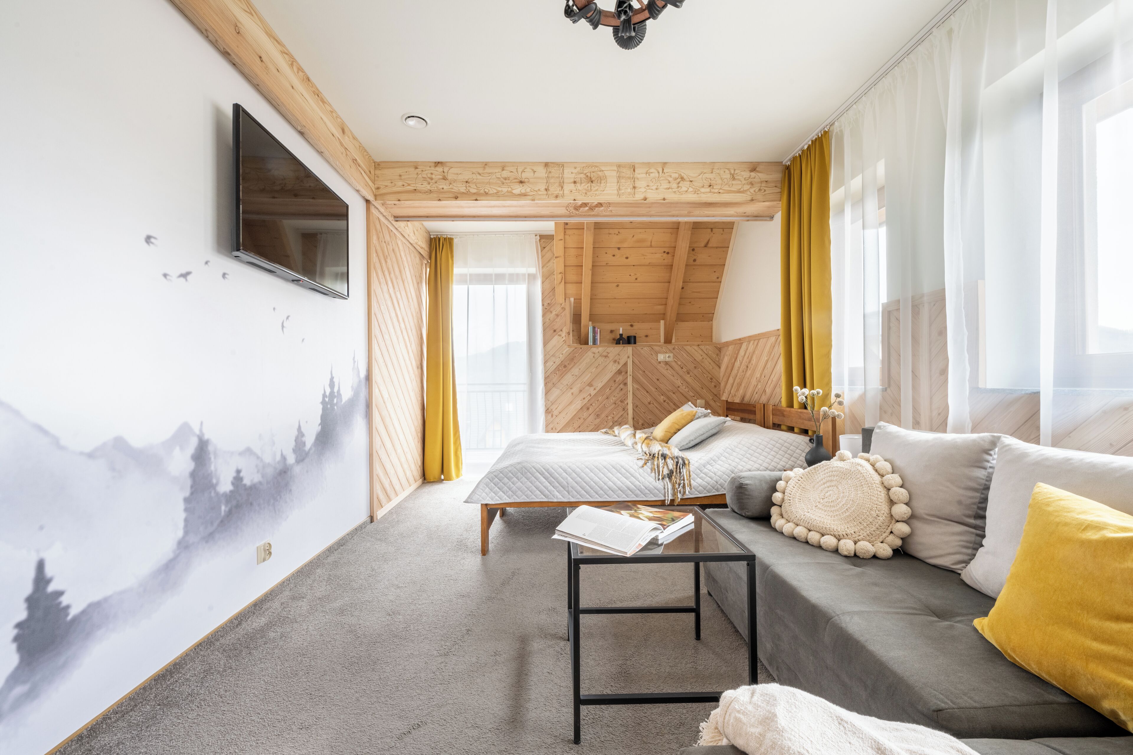A modern bedroom with a sloped ceiling, a bed with a grey duvet, a sofa with yellow cushions, and a wall-mounted TV.