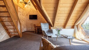 Acogedor dormitorio en el ático con vigas de madera, una araña, un sillón gris, un sofá y una mesita con un jarrón de flores.