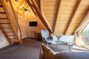 Acogedor dormitorio en el ático con vigas de madera, una araña, un sillón gris, un sofá y una mesita con un jarrón de flores.
