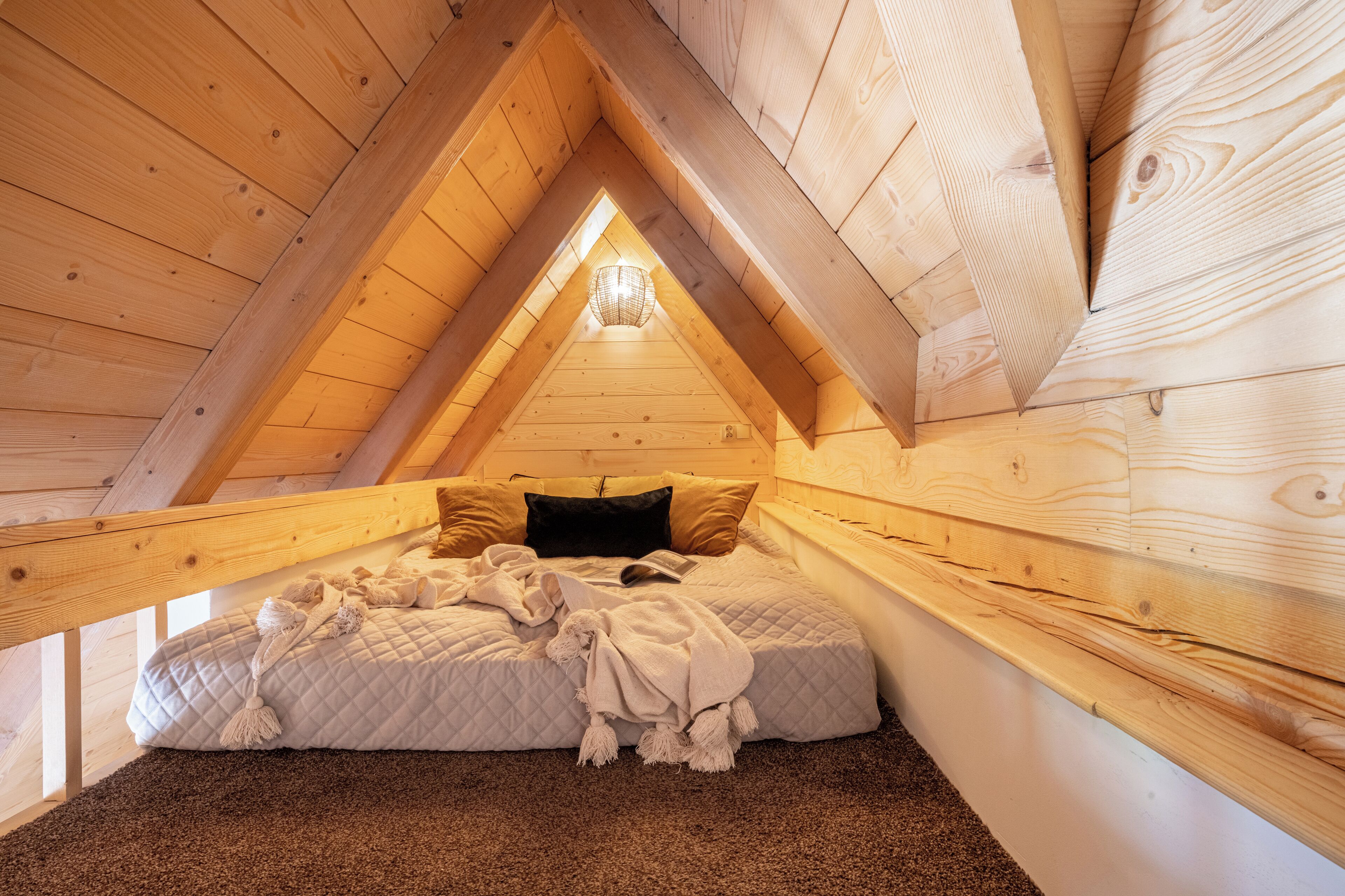 A cozy attic bedroom with a sloped ceiling, wooden beams, a bed with a quilted blanket, and a small window.