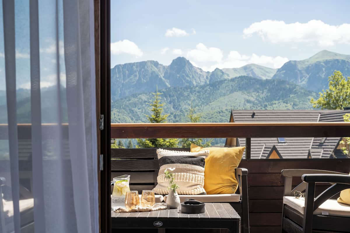 Un balcon avec un banc en bois, une table avec des boissons et un vase, et une vue sur les montagnes.