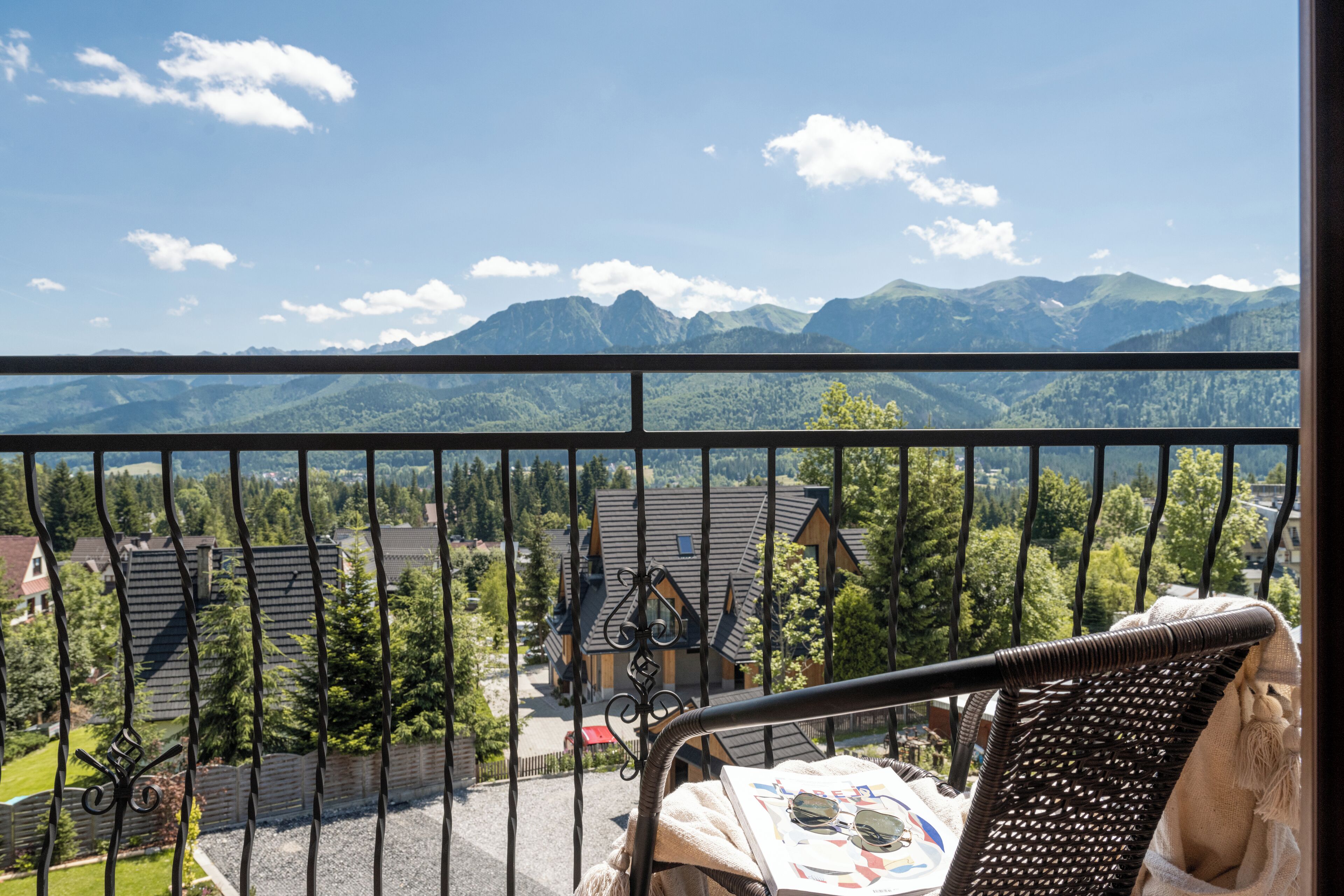 A balcony with a view of mountains, a chair, and a table with a newspaper and sunglasses.