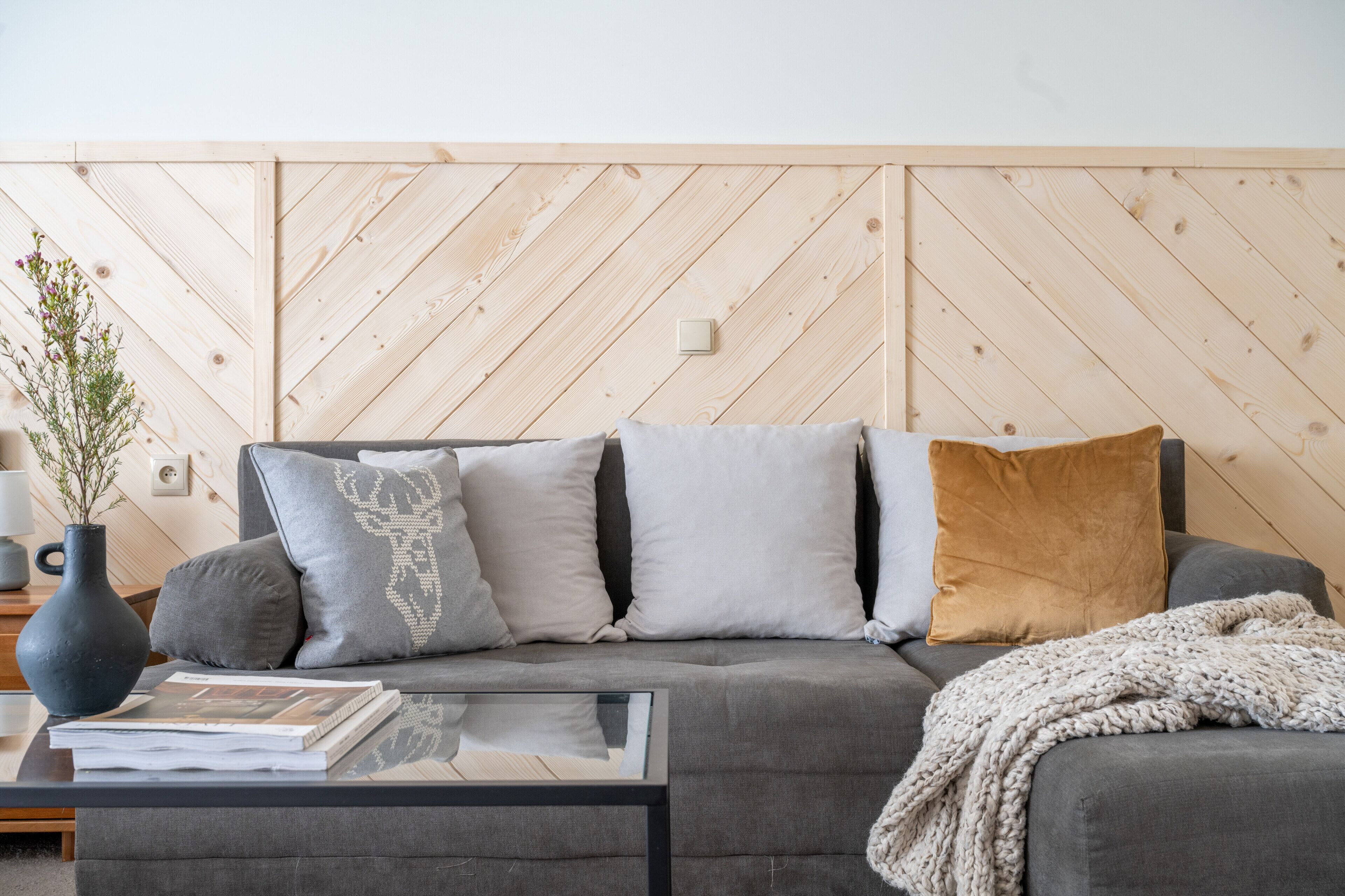 A modern living room with a grey sofa, multiple cushions, a glass coffee table, and a wooden wall panel.