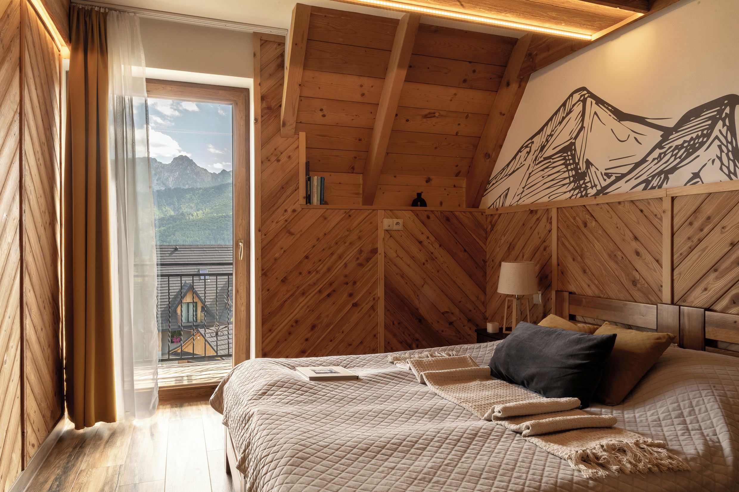Un dormitorio con techo de madera, una cama con cubrecama acolchado, una mesita de noche con lámpara y vistas a montañas a través de una ventana.