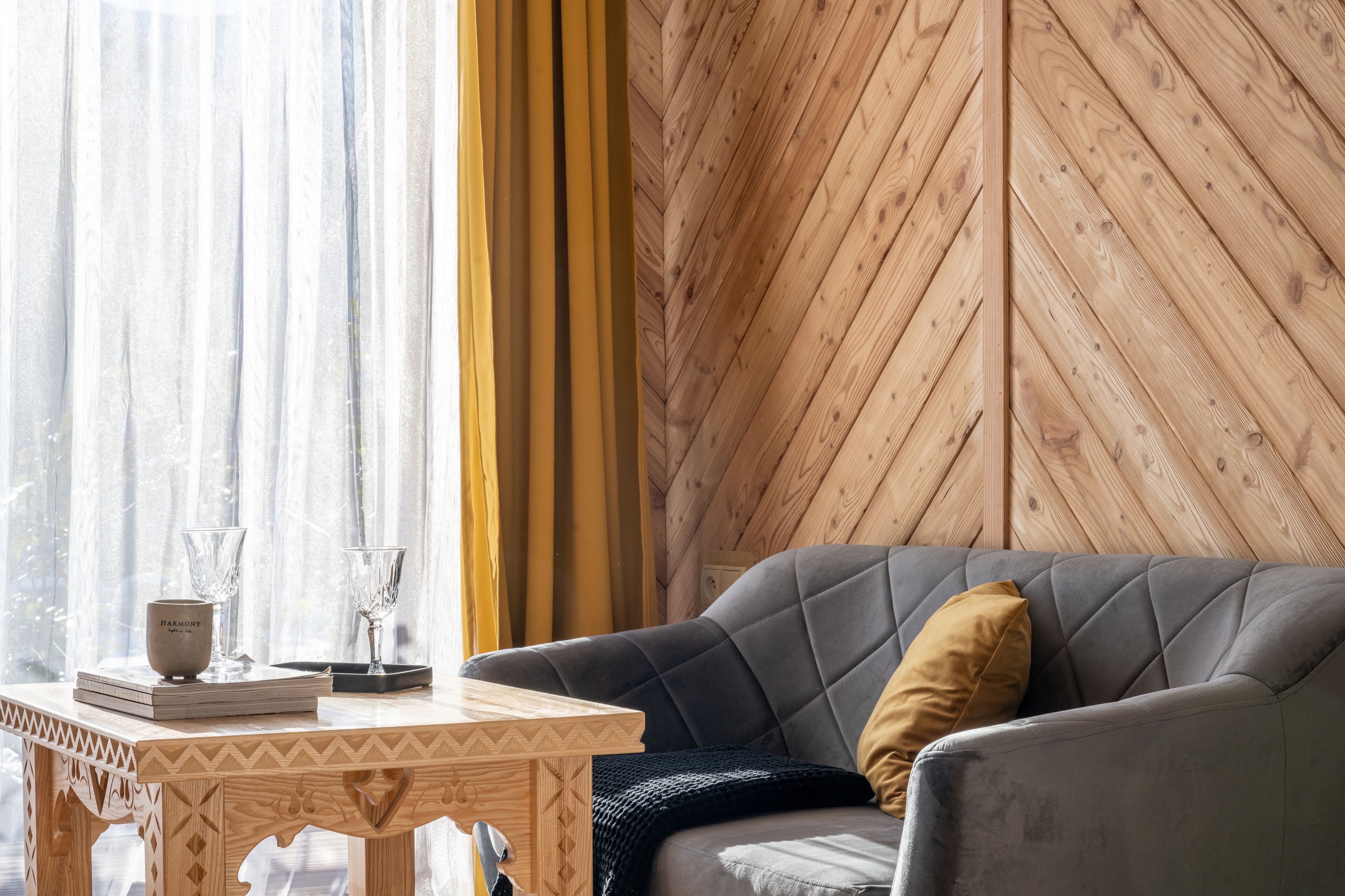 A cozy living room with a grey sofa, a wooden table with a patterned top, and a yellow curtain.