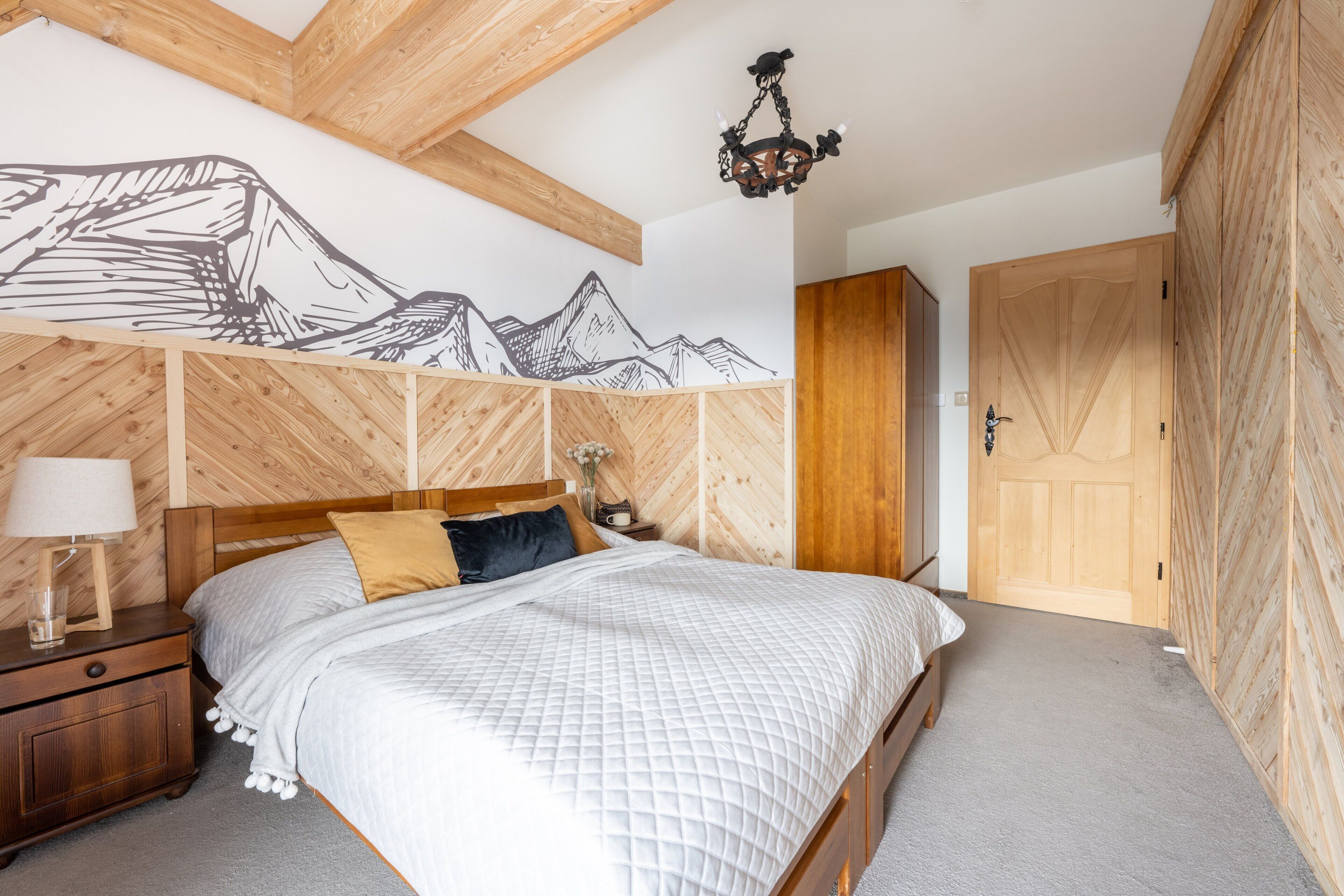 Une chambre à coucher avec une tête de lit en bois, un lit recouvert d’une couette blanche matelassée, une table de chevet avec une lampe et un panneau en bois fixé au mur, orné d’un motif de montagne.
