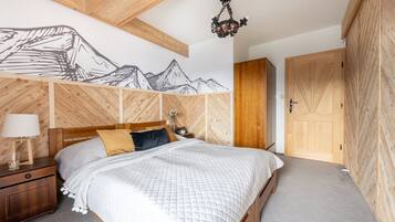 A bedroom with a wooden headboard, a bed with a quilted white duvet, a nightstand with a lamp, and a wall-mounted wooden panel with a mountain design.