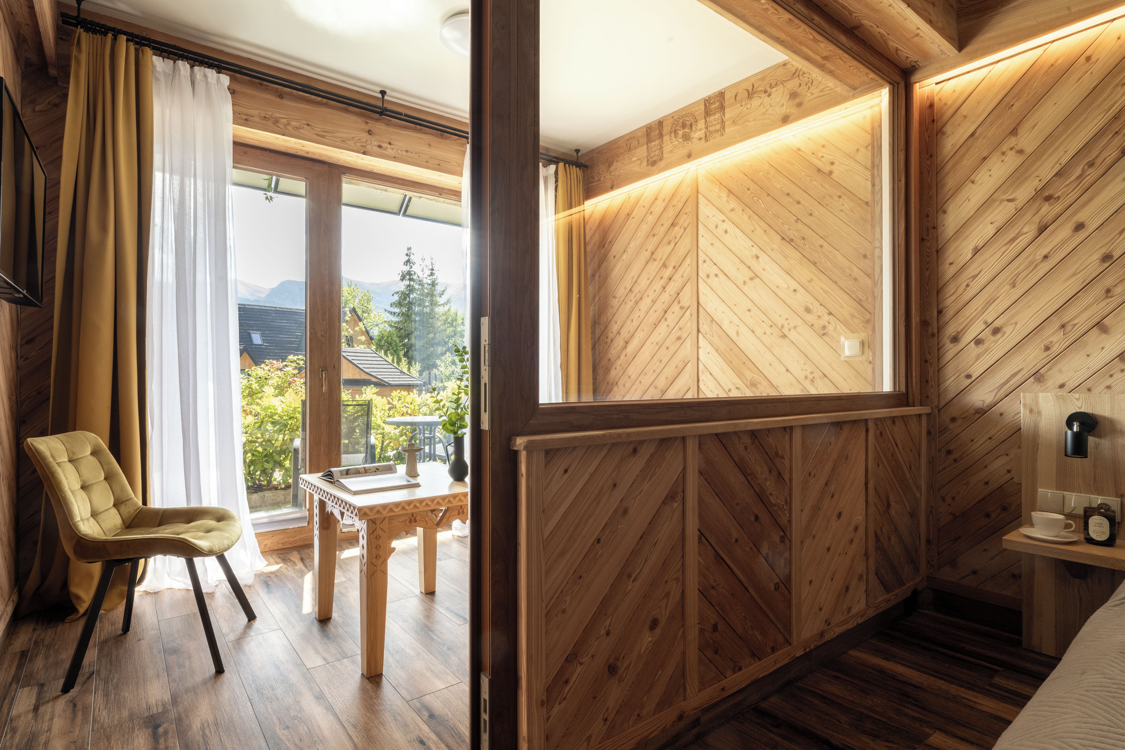 Una habitación con suelo de madera, una silla, una mesita y vistas a zonas verdes y montañas a través de una ventana.