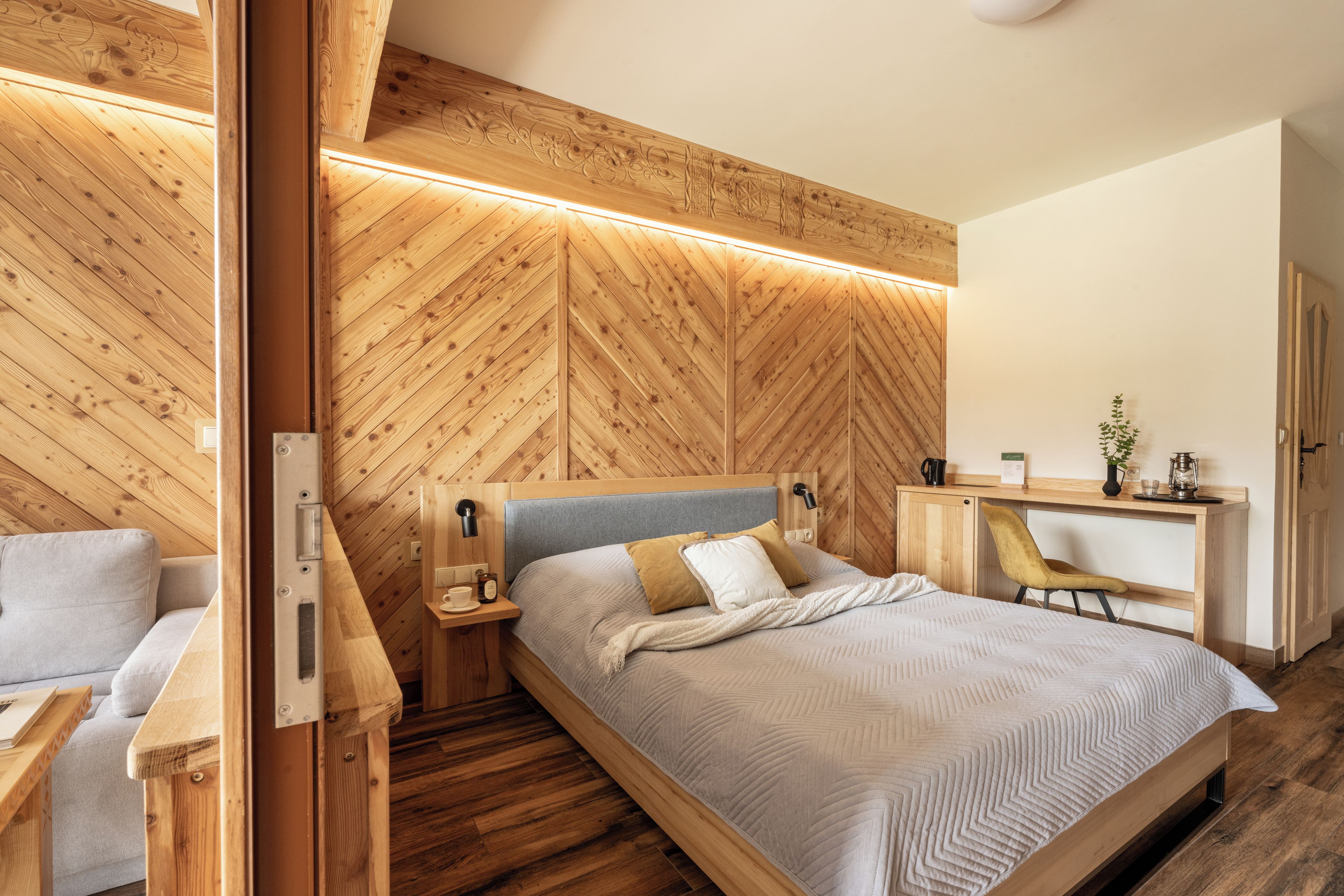 Une chambre moderne avec un mur en bois à motifs en chevrons, un lit avec une tête de lit grise, une table de chevet avec une lampe et un bureau avec une chaise.