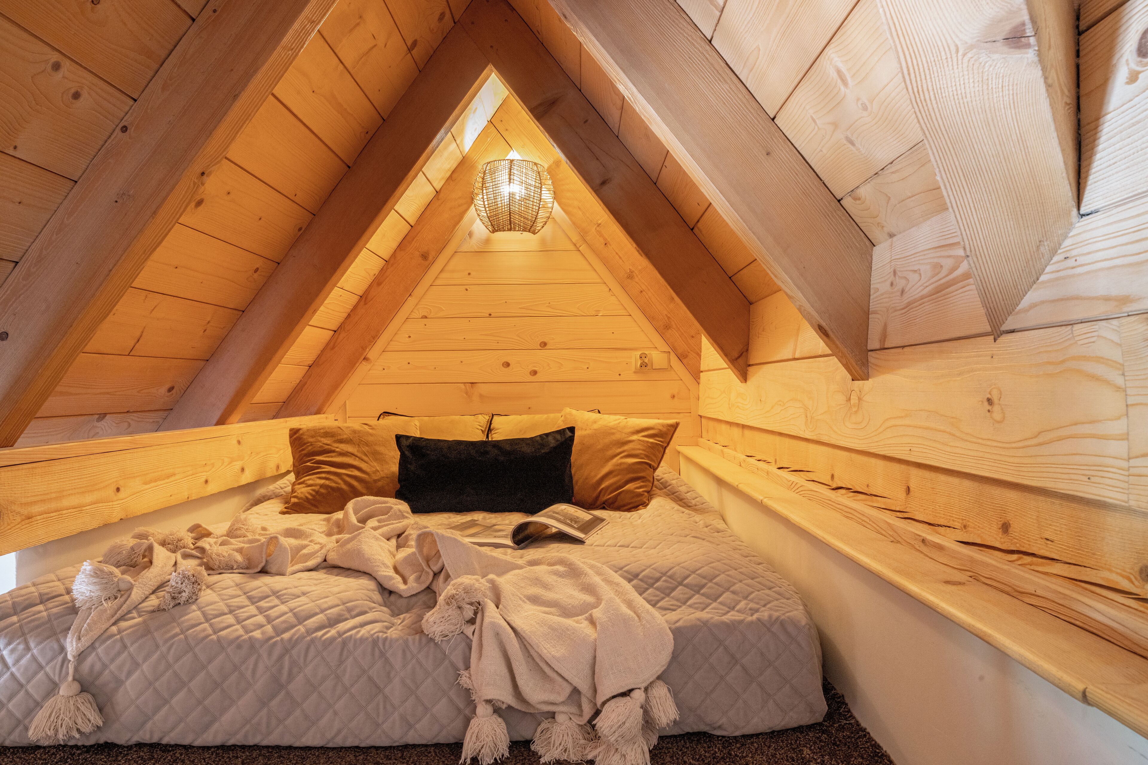 Une chambre mansardée chaleureuse, avec un plafond en pente en bois, un lit recouvert d’une courtepointe et un luminaire suspendu.