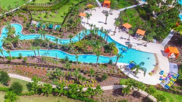 An aerial view of a resort with a winding pool, surrounded by palm trees and a small beach area with umbrellas. - Last Minute Disney Getaway! Private Pool & Themed Bedrooms. Book Now! #81801 (Orlando)