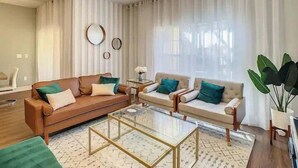 Uma sala de estar moderna com um sofá marrom, duas poltronas, uma mesa de centro de vidro e uma janela grande com cortinas leves.