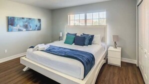 Um quarto com uma cama, uma mesinha de cabeceira, uma janela e um quadro na parede.