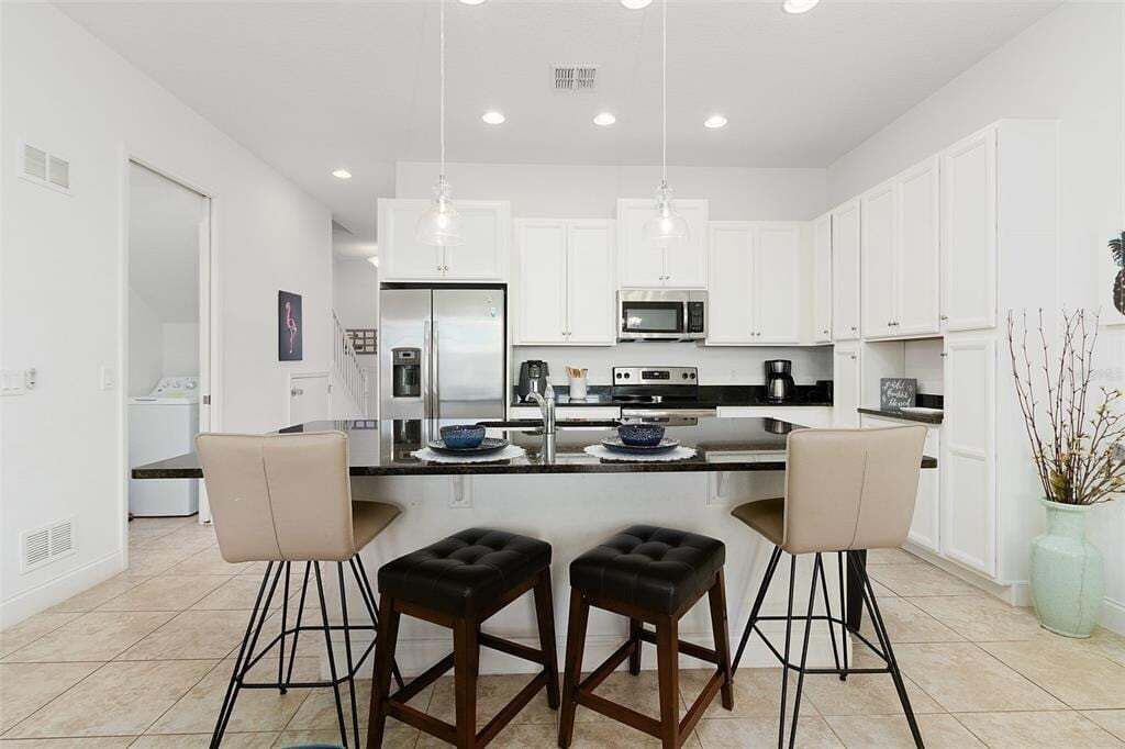 A modern kitchen with white cabinets, a central island with stools, a refrigerator, microwave, and a sink.