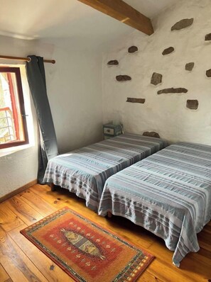 A small, cozy room with two beds, a wooden floor, a stone wall, and a window with curtains.