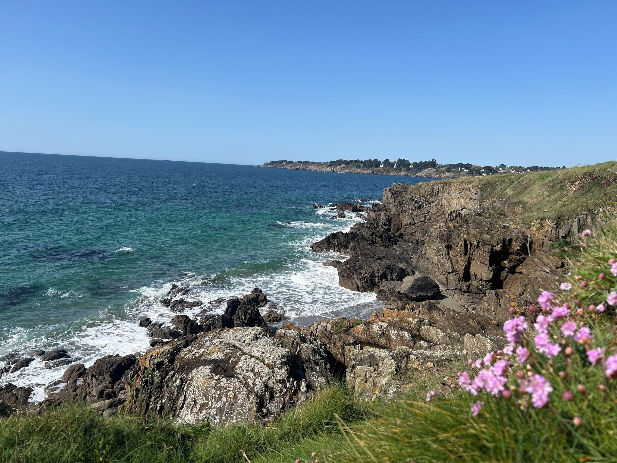 Paisagem costeira com falésias rochosas, mar azul límpido e edifícios ao longe.