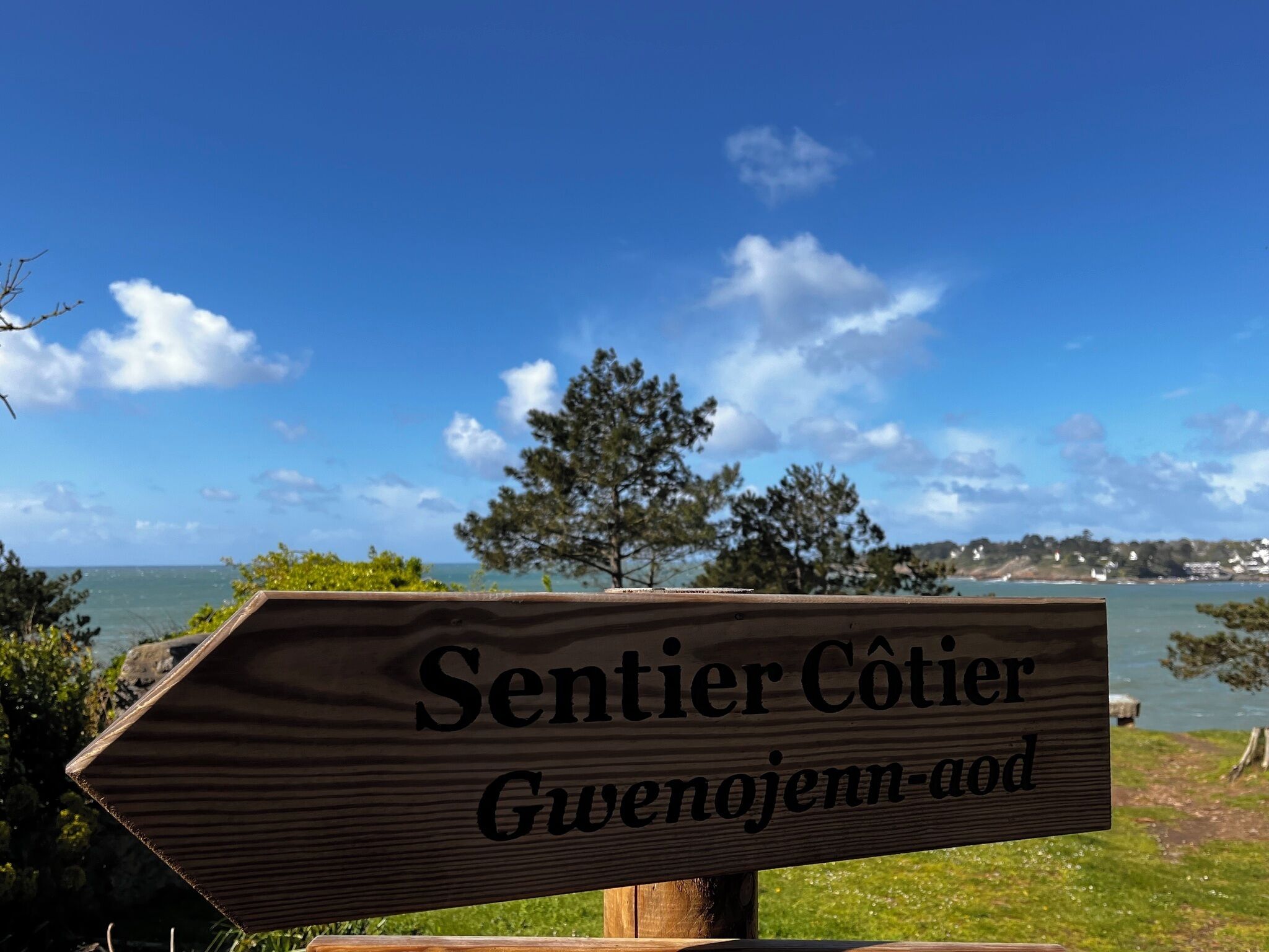 Un letrero de madera con el texto 'Sentier Côtier Gwentjenn-aod' sobre un cielo azul despejado, un cuerpo de agua y una costa distante con árboles y edificios.