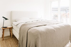 A neatly made bed with white linens and a beige blanket, a bedside table with a lamp, and a large window providing a view of the outside.