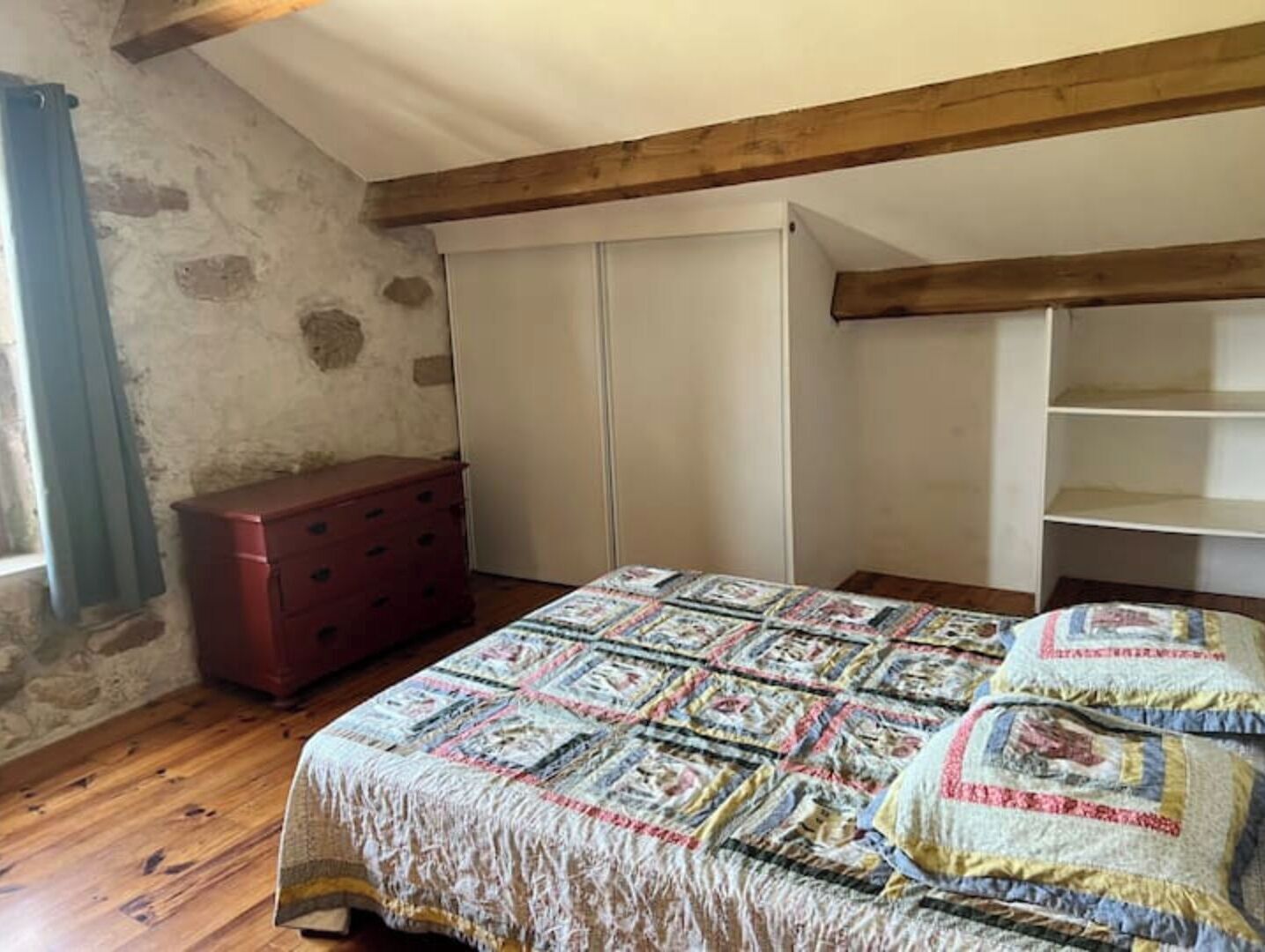 A bedroom with a wooden floor, a bed with a patterned quilt, a red dresser, and a white wardrobe.