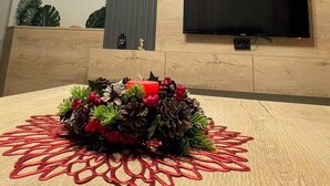 A modern living room with a flat-screen TV mounted on a wooden wall, a decorative wall hanging, and a table with a floral centerpiece. - Nesi apartment  (Pristina)
