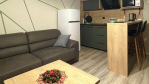 A modern living space with a sofa, a small wooden table with a floral centerpiece, a kitchen area with a microwave and kettle, and a unique geometric wall design. - Nesi apartment  (Pristina)