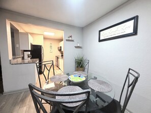 A dining area with a glass table surrounded by chairs, a kitchen in the background, and a motivational quote on the wall.