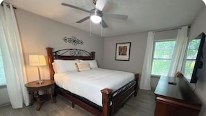 Quarto com cama de madeira, mesa de cabeceira, escrivaninha, ventoinha no teto e janela com cortinas.