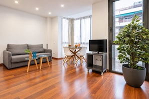 Ein modernes Wohnzimmer mit einem grauen Sofa, einem kleinen Tisch mit einer Pflanze, einem Essbereich mit Fernseher und einer großen Kübelpflanze.