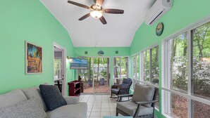 Un salon avec un mur vert, un ventilateur de plafond, un canapé, un fauteuil et de grandes fenêtres donnant sur des arbres et une terrasse.