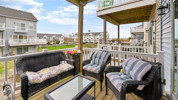 A covered outdoor seating area with wicker furniture and a glass table, overlooking a residential neighborhood. - Seaside Village 4 by Seaside Vacations & Sales (Ocean City)