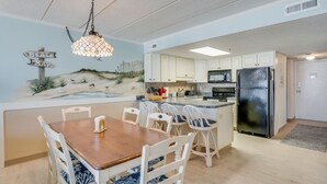 Una cocina con gabinetes blancos, un refrigerador negro y una zona de comedor con una mesa de madera y sillas blancas.