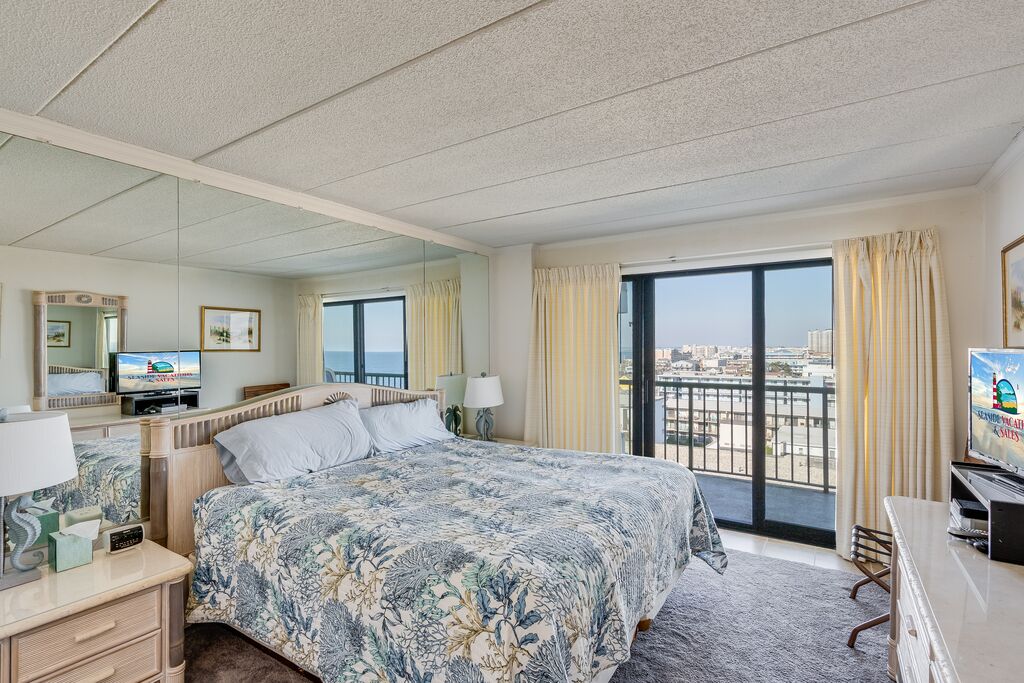 Ein Hotelzimmer mit einem großen Bett, einem Fernseher, einem Schreibtisch und einem Balkon mit Blick auf die Stadt.