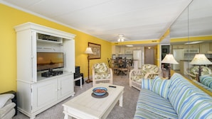 A living room with a TV, white cabinets, a striped sofa, and a dining area with chairs and a table. - Summer Beach 807 by Seaside Vacations & Sales (Ocean City)