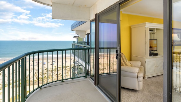 A balcony with a view of the beach and ocean, a sliding glass door leading to a room with a TV and a cabinet. - Summer Beach 807 by Seaside Vacations & Sales (Ocean City)