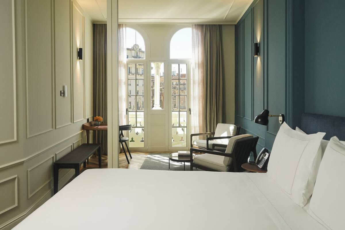 A hotel room with a large bed, a desk with a chair, a sofa, and a view of the city through a glass door.