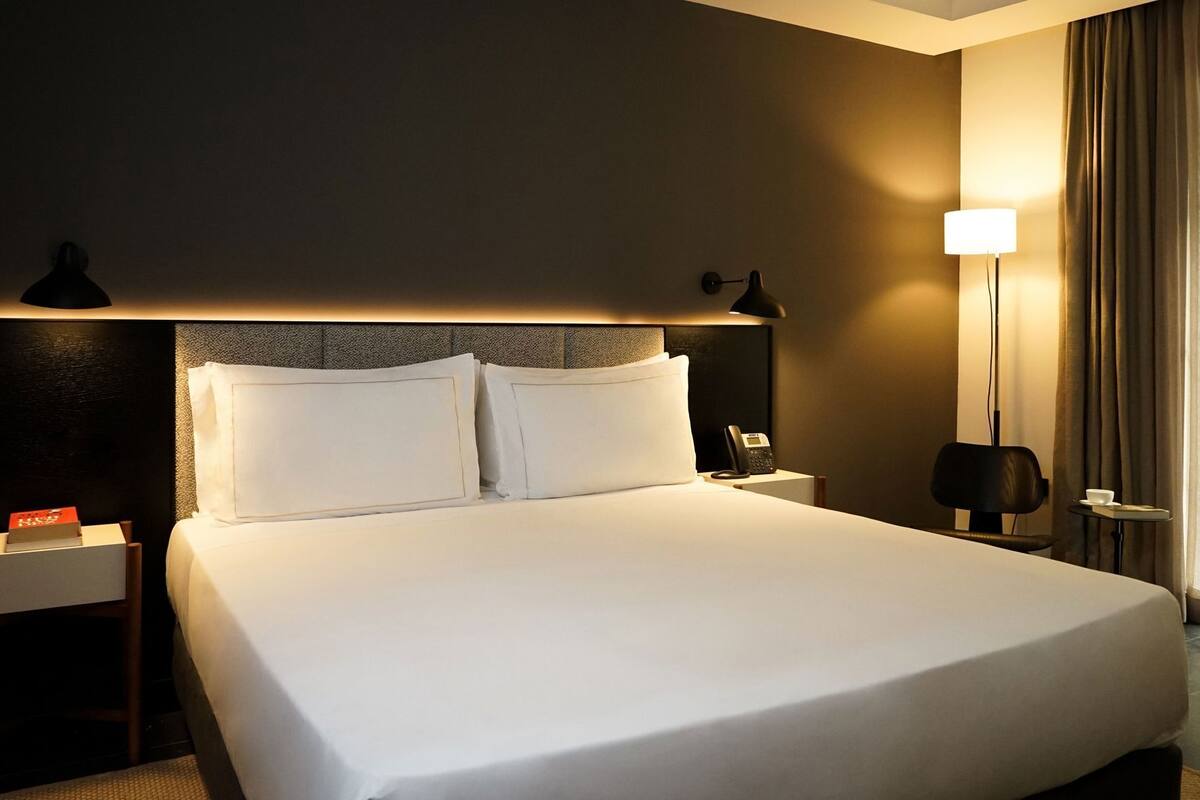 A hotel room with a neatly made bed, a bedside phone, a lamp, and a small table with a cup.