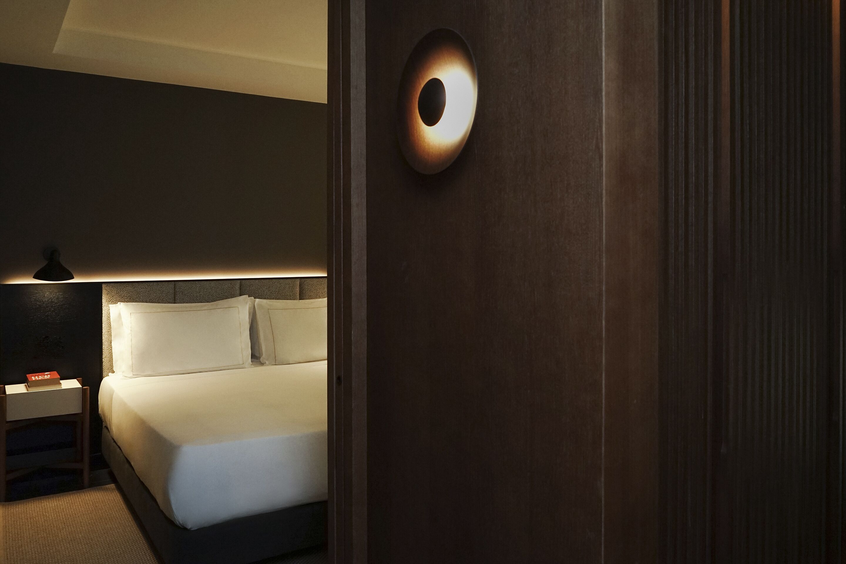 Una habitación de hotel moderna con una cama bien hecha, una mesita de noche con lámpara y una lámpara de pared.