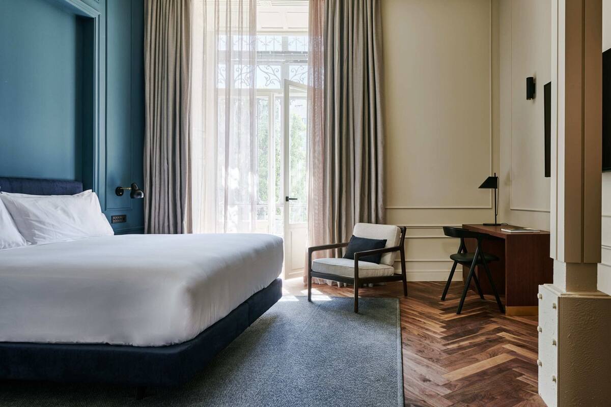 A hotel room with a large bed, a chair, a desk, and a view of greenery through the window.