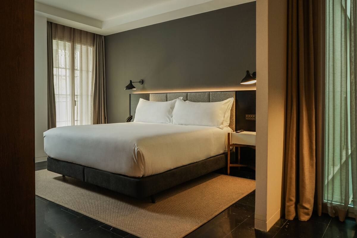 A modern hotel room with a large bed, a bedside table, a lamp, and a view of the outdoors.