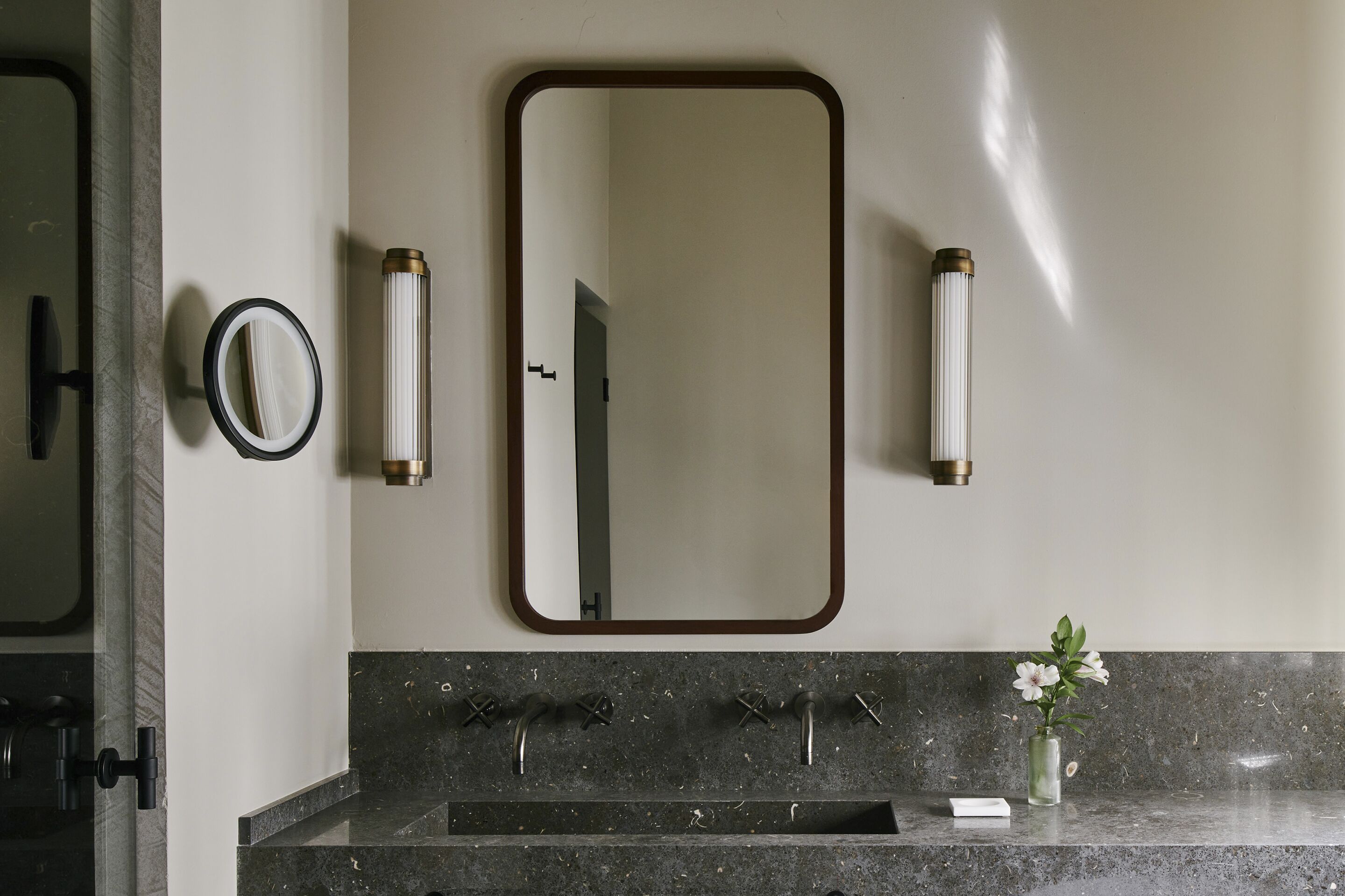 Un baño con un espejo grande, dos dispensadores de toallas en la pared, un lavabo con tres grifos y un florero con flores en la encimera.