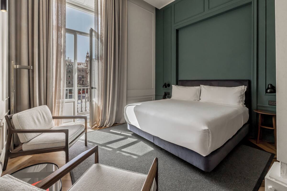 A hotel room with a large bed, a chair, a view of the city through the window, and a green accent wall.