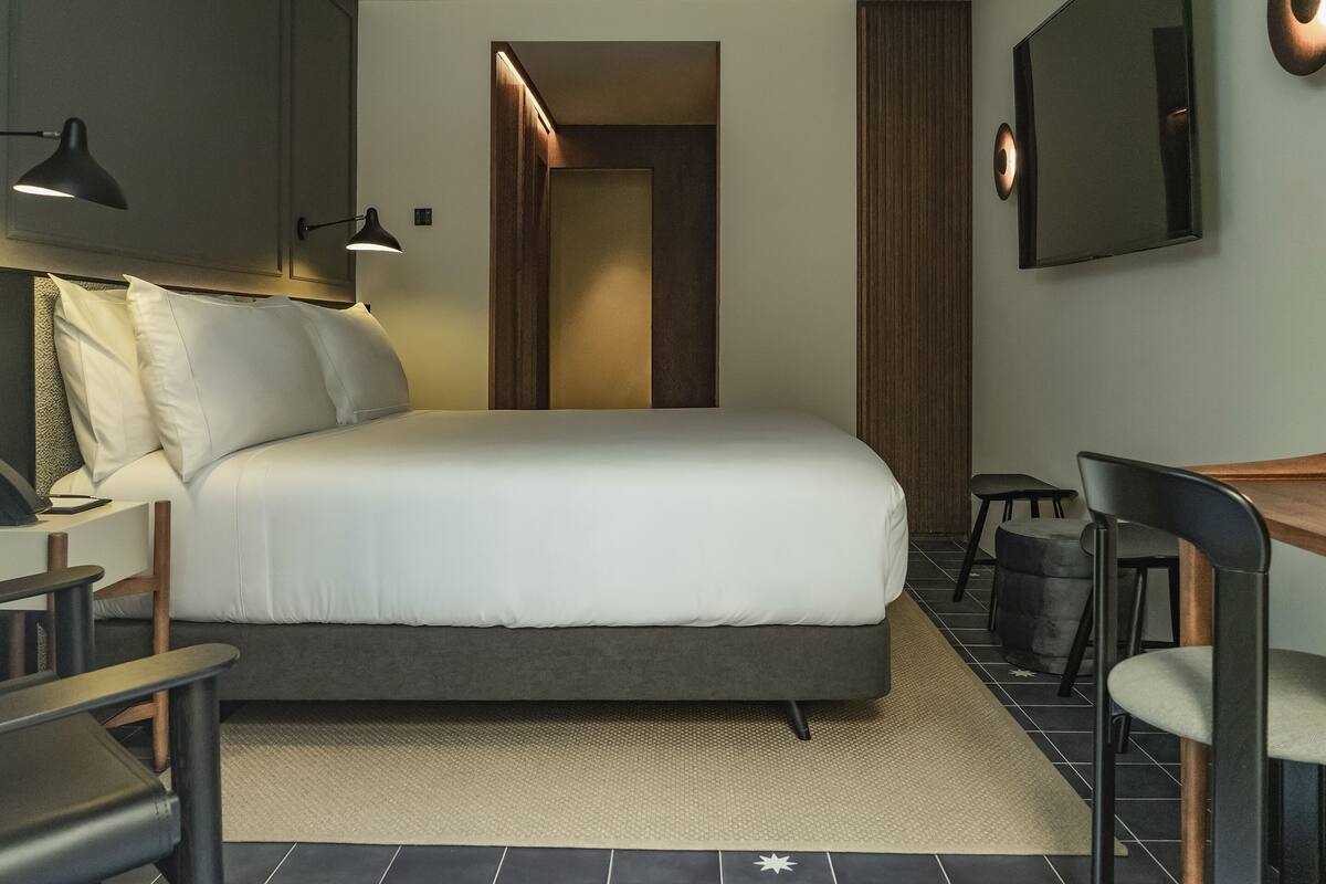 A modern hotel room with a large bed, a desk, and a chair. There are two wall-mounted lamps and a mirror.