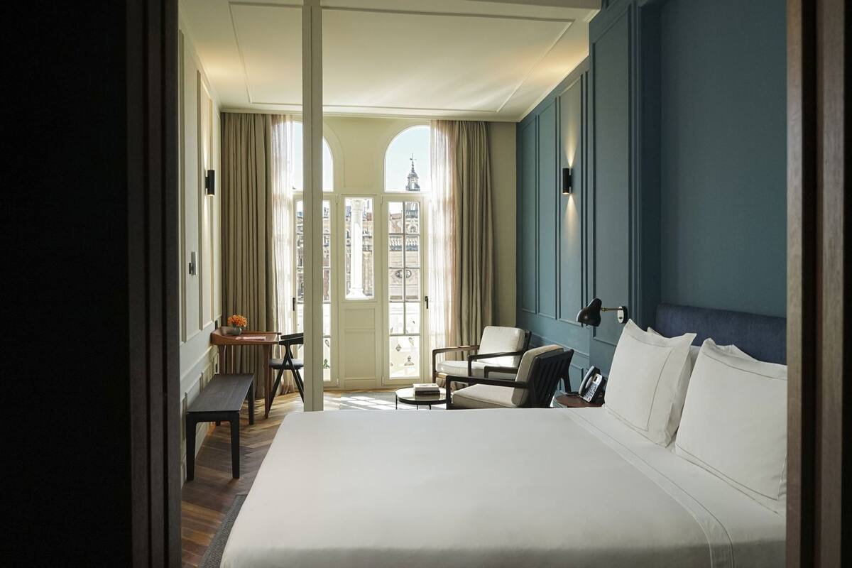 A hotel room with a large bed, a desk with a chair, a small table, and a sofa. There is a window with curtains and a view of a building outside.