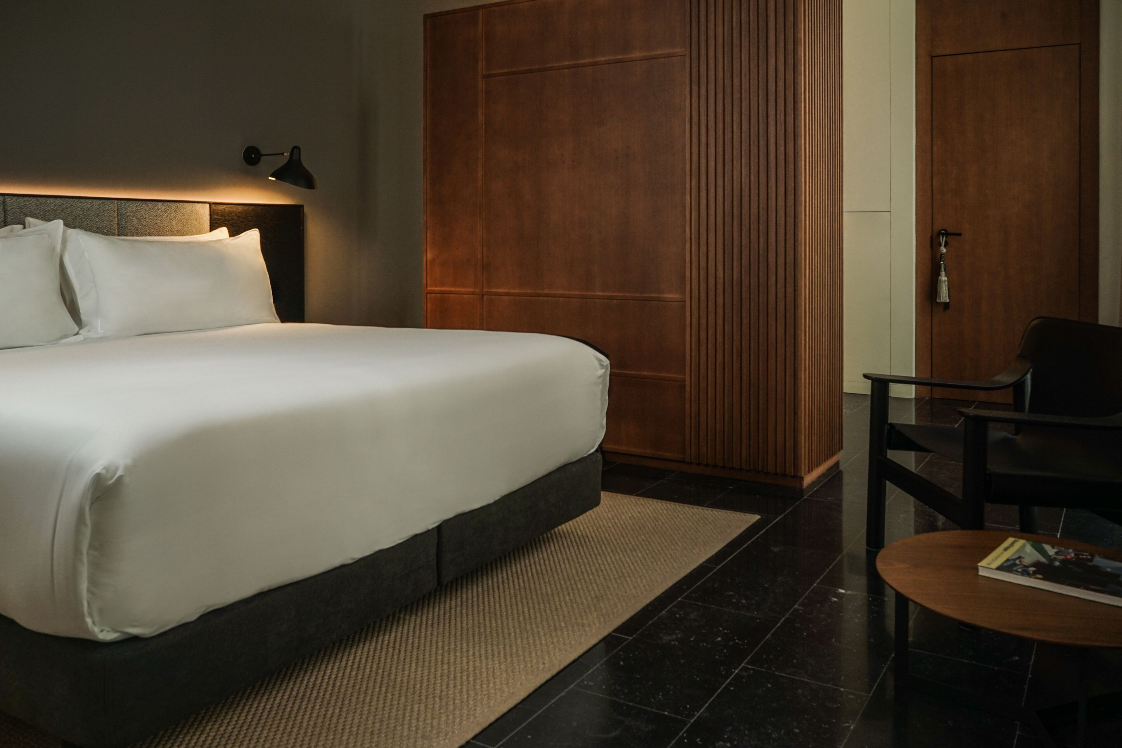 A modern hotel room with a large bed, a wooden wardrobe, a small table, and a desk.