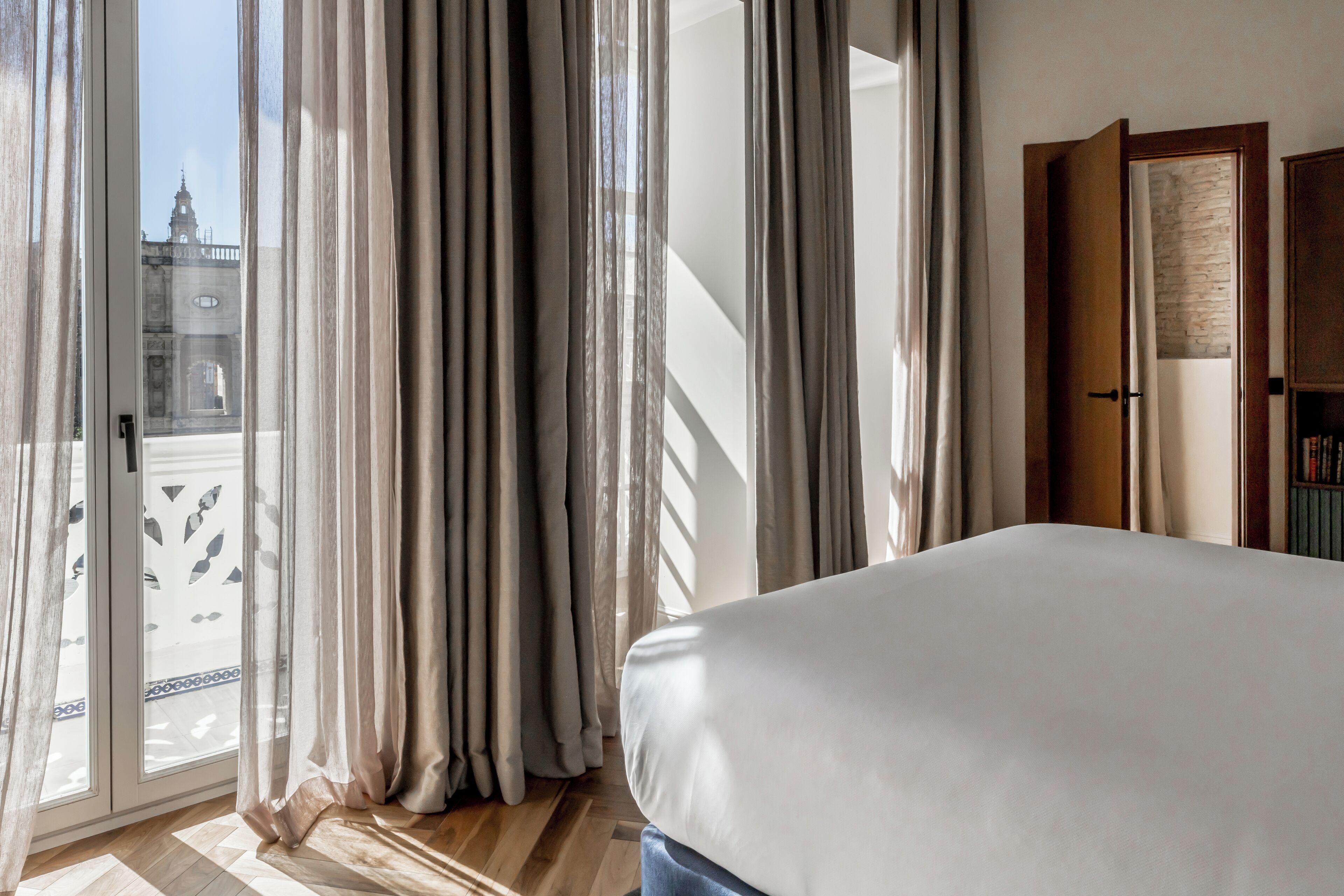 Ein Schlafzimmer mit einem großen Bett, einer Glastür, die auf einen Balkon führt, und einer Holztür rechts.