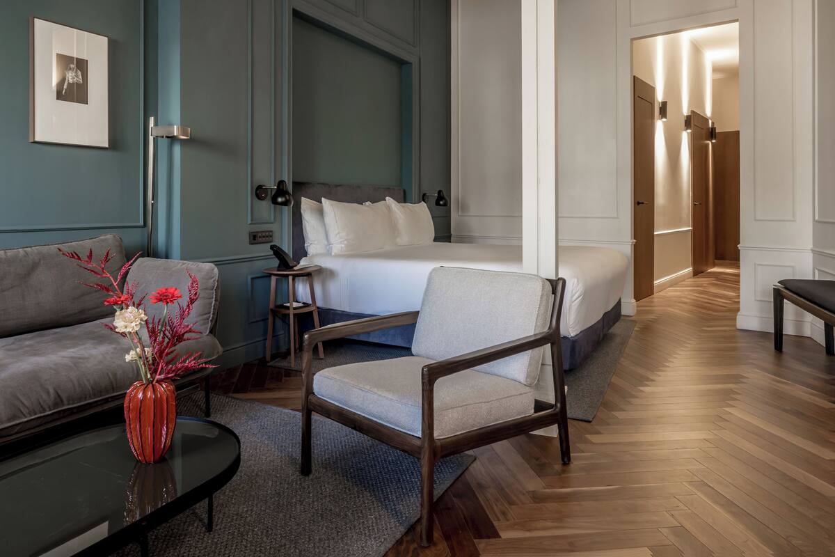 A modern hotel room with a bed, a sofa, a chair, a small table, and a vase with flowers.
