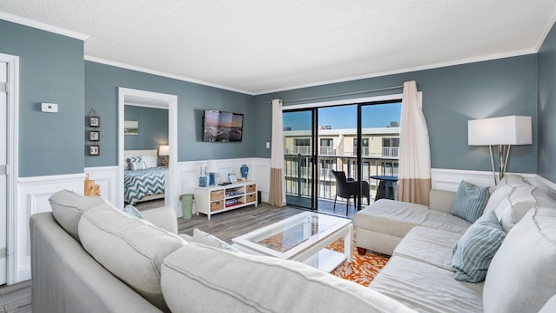 A modern living room with a sofa, a coffee table, and a TV. There is a balcony with a view of buildings. - Wight Bay 559 by Seaside Vacations & Sales (Ocean City)